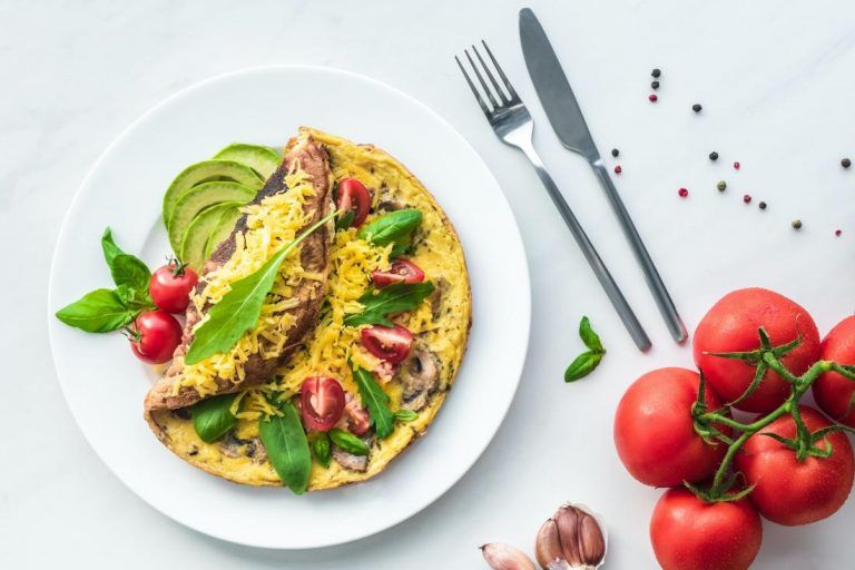 flat lay with omelette with cherry tomatoes, avocado pieces and cutlery on white marble surface