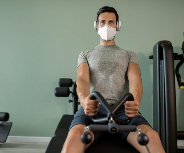 Hombre atlético con mascarilla con entrenamiento deportivo en una máquina de remo en un gimnasio.