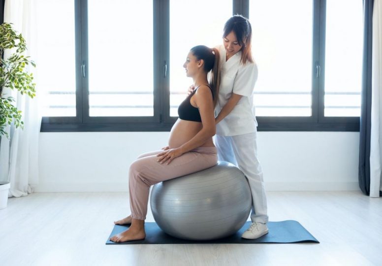 Fisioterapeuta ayudando a embarazadas a hacer ejercicios con pelota preparándose para el parto en casa.